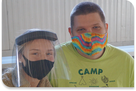 Two people in covid masks, one with a clear face screen and one with a colorful purple, orange, and green mask
